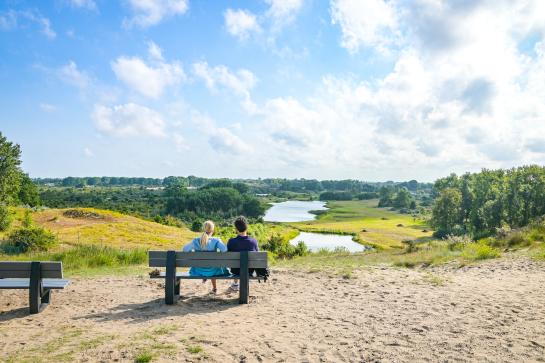 Een koppel rust uit op een bankje met zicht op een duinenlandschap